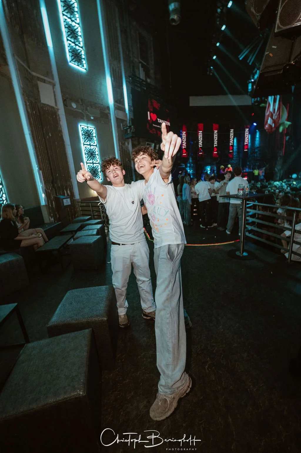 Two young men are standing in a dance club, posing for a photo. In the background are other people and illuminated signs reading "I Love Dancing."