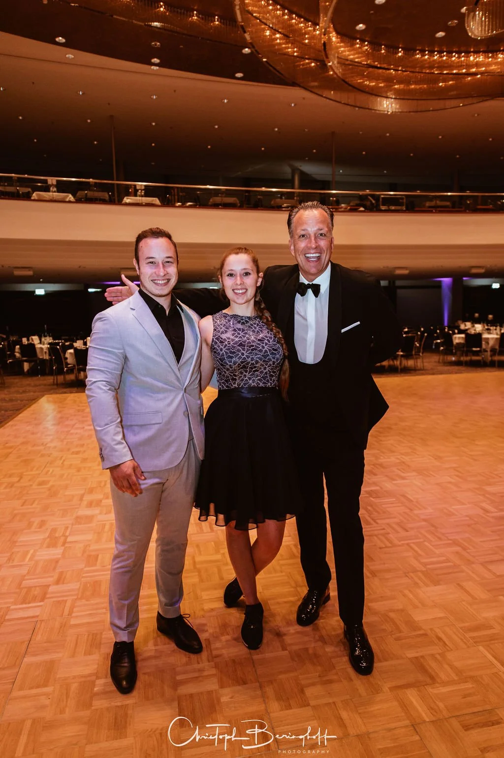 Drei Menschen in festlicher Kleidung auf einer Tanzfläche bei einem Event, die beiden Männer tragen Anzüge, die Frau trägt ein Kleid, alle lachen und schauen in die Kamera.