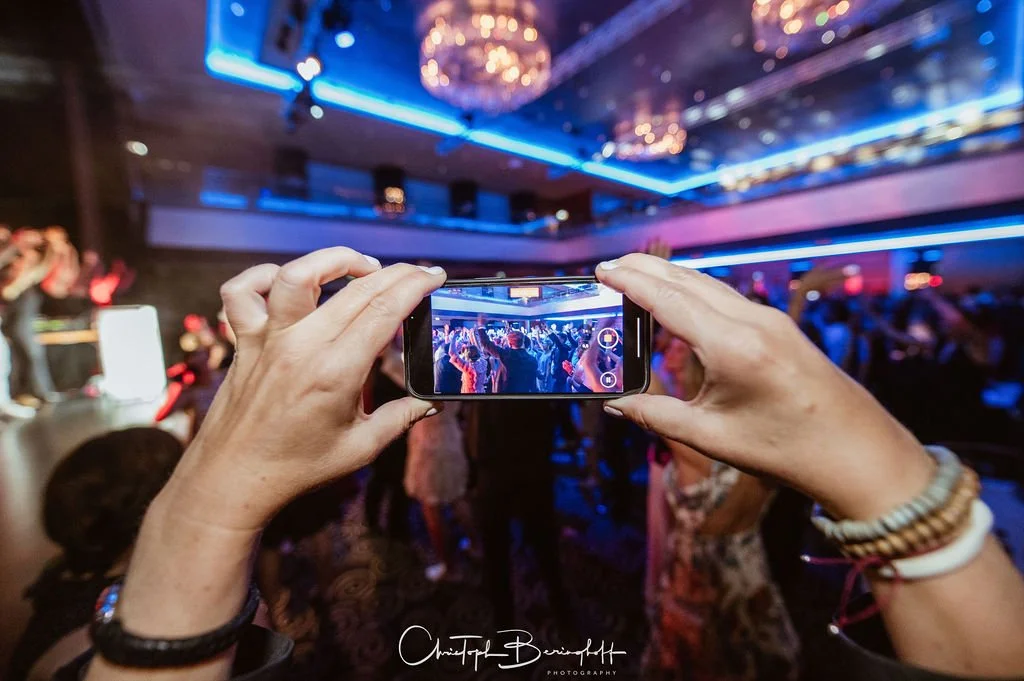 Person fotografiert eine Bühne bei einer Veranstaltung mit einem Smartphone, im Hintergrund ein Publikum und bunte Lichter.