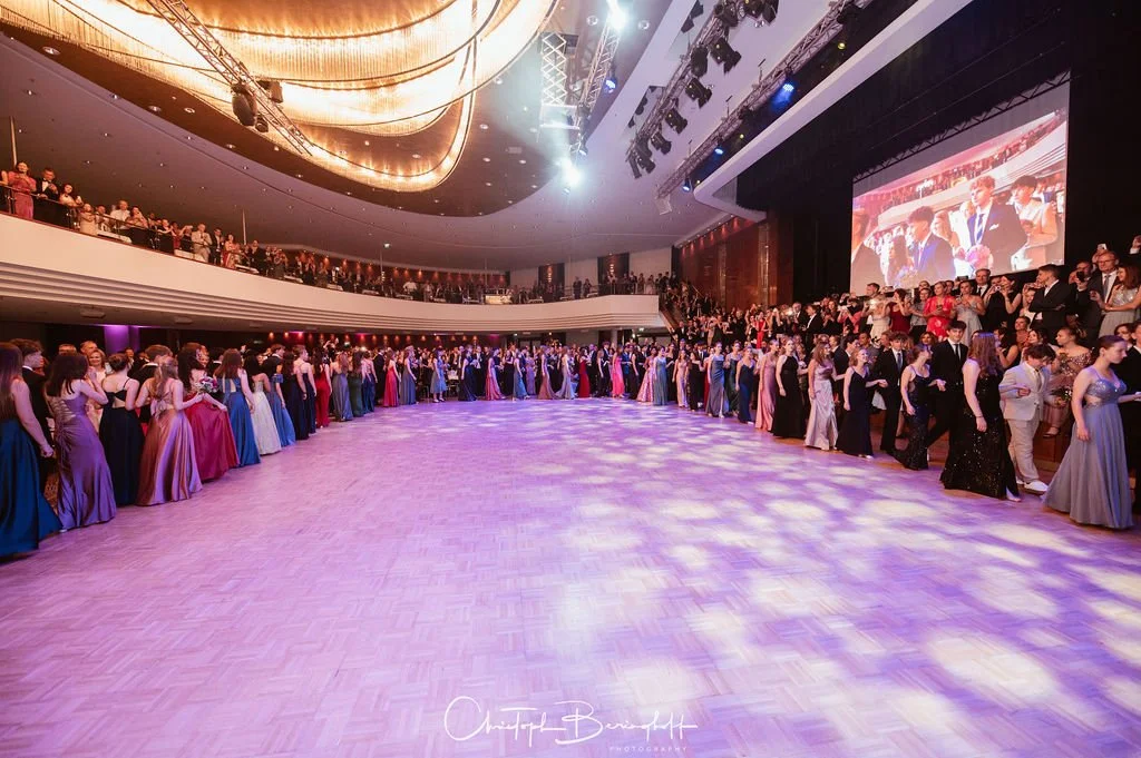 Tanzball mit vielen jungen Leuten in formeller Kleidung, im Saal mit Tanzfläche und großer Bühne mit Leinwand, dekoriert mit Lichtern und moderner Beleuchtung.