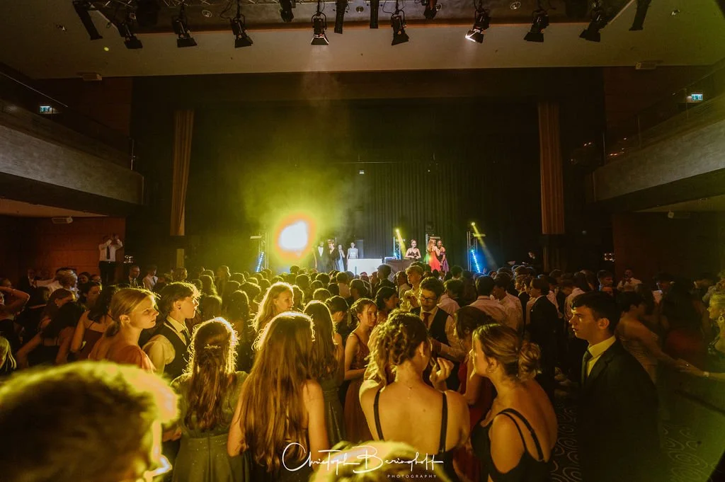 Menschen tanzen auf einer Party vor einer Bühne mit einem DJ und Sängerinnen, bunte Lichteffekte auf der Tanzfläche