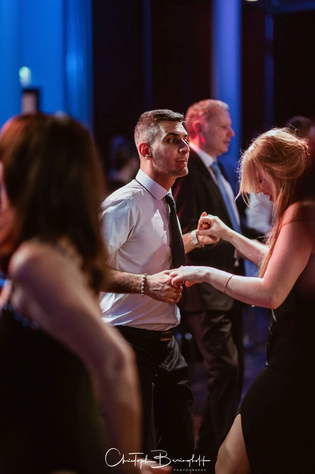 Menschen tanzen auf einer Party im dunklen Raum, einige halten sich an den Händen, im Vordergrund ein Mann mit weißem Hemd und schwarzer Krawatte, im Hintergrund weitere Tänzer.