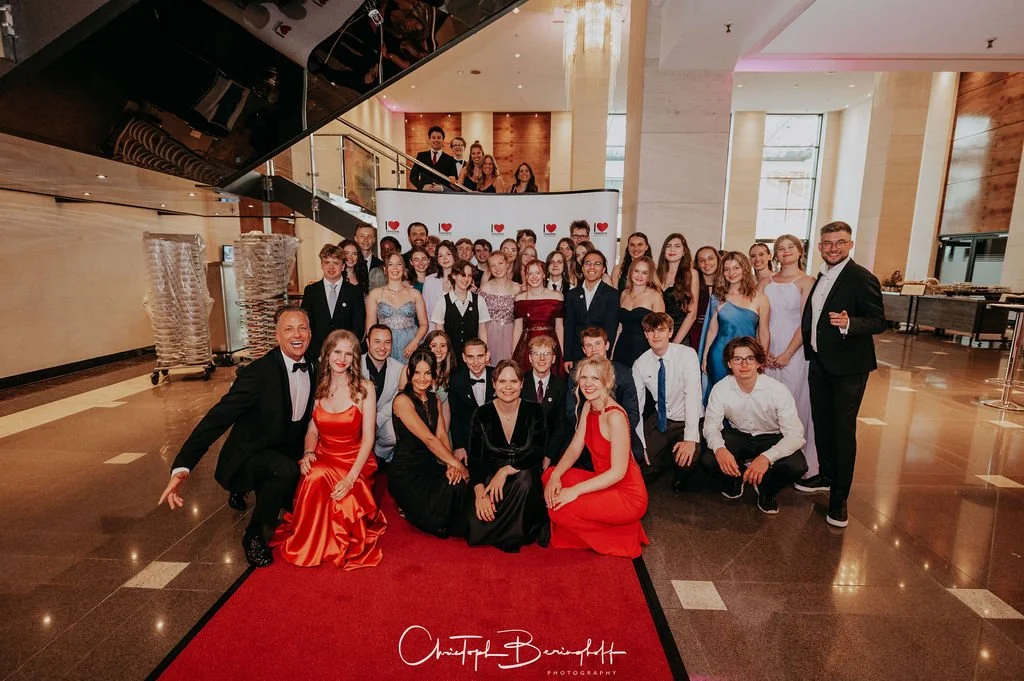 Gruppenfoto von jungen Menschen in formeller Kleidung bei einer Gala oder Feier in einem festlich dekorierten Innenraum.