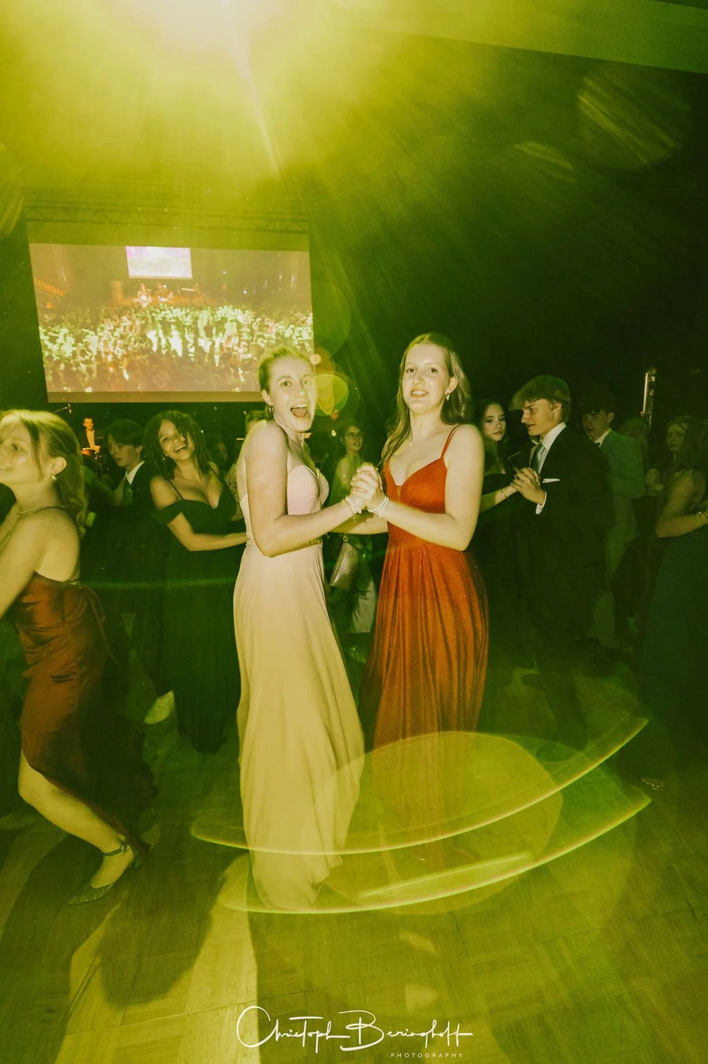Zwei Frauen tanzen auf einer Party, während im Hintergrund weitere Gäste tanzen und eine große Leinwand eine Menschenmenge zeigt.