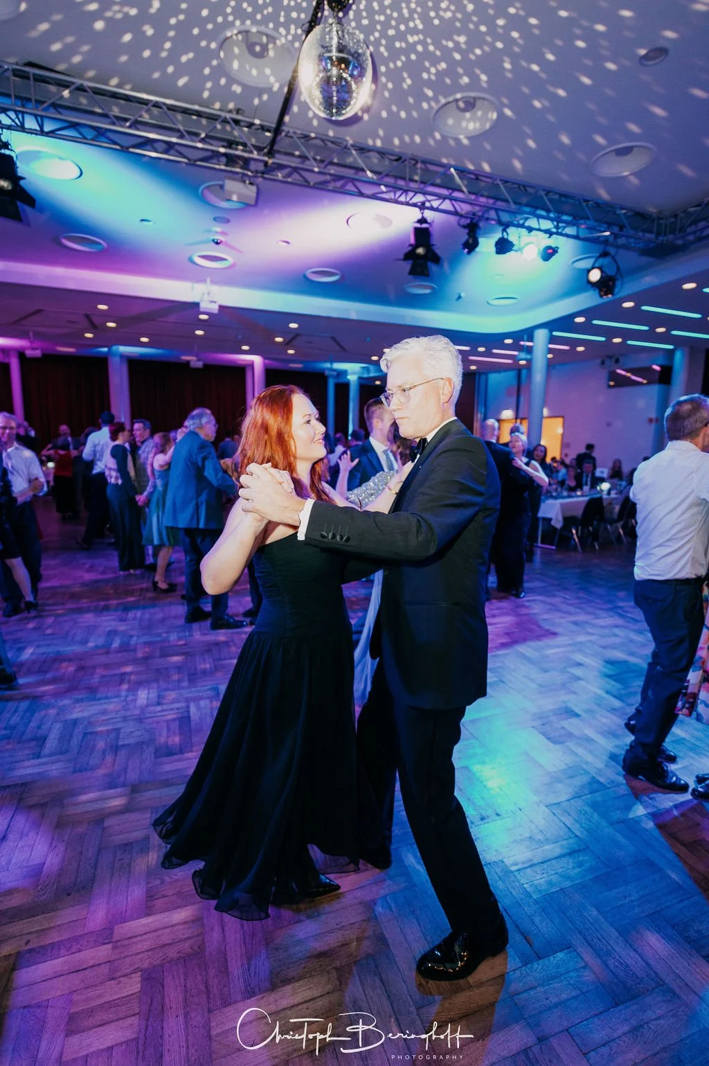 Paare tanzen auf einer Feier in einem festlich beleuchteten Raum mit Disco-Kugel an der Decke, die Lichtreflexe auf die Decke wirft, während andere Gäste im Hintergrund sitzen und sprechen.