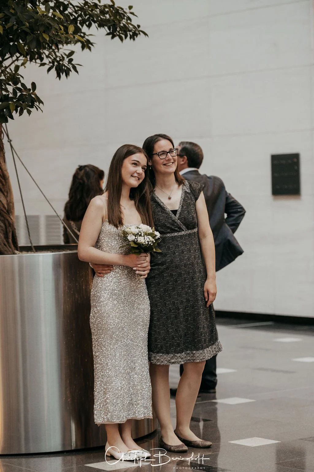 Zwei Frauen, eine in einem funkelnden Kleid mit Blumenstrauß, die andere in einem schwarzen Kleid mit Mustern, stehen gemeinsam und lächeln bei einer Veranstaltung.