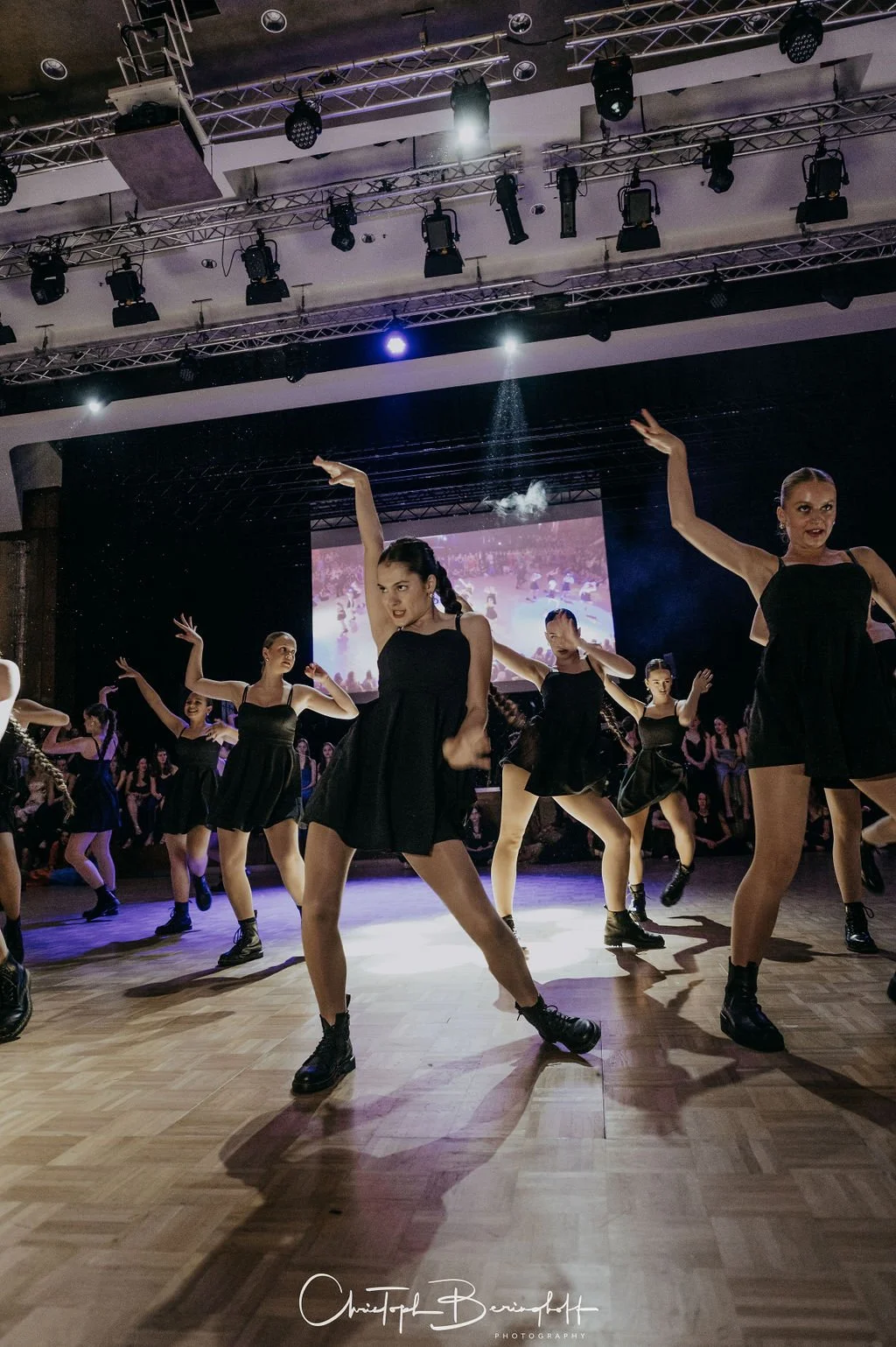 Tanzgruppe auf einer Bühne während eines Auftritts, alle tragen schwarze Kleider und schwarze Stiefel, mit Scheinwerfern und Leinwand im Hintergrund.