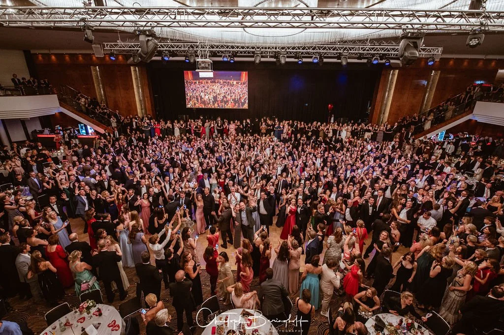 Viele Menschen tanzen und feiern in einem großen Festsaal bei einer Party, umgeben von Tischen mit gedeckten Plätzen.