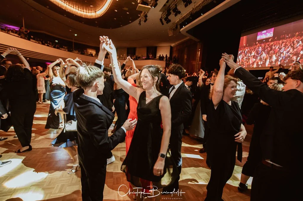 Menschen tanzen bei einer festlichen Veranstaltung in einem großen Saal, überwiegend in eleganter Kleidung, mit einem großen Bildschirm im Hintergrund.