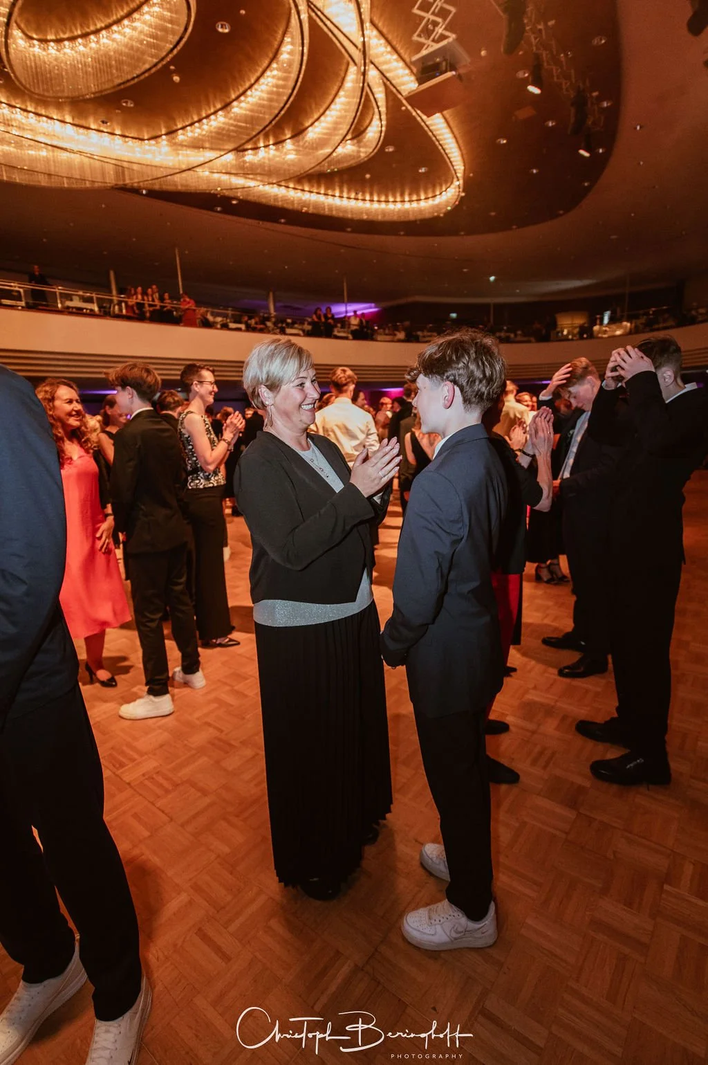 Eine Frau und ein junger Mann tanzen und unterhalten sich in einem festlichen Raum mit hoher Decke und warmem Licht. Im Hintergrund sind weitere Menschen, die ebenfalls feiern oder tanzen.