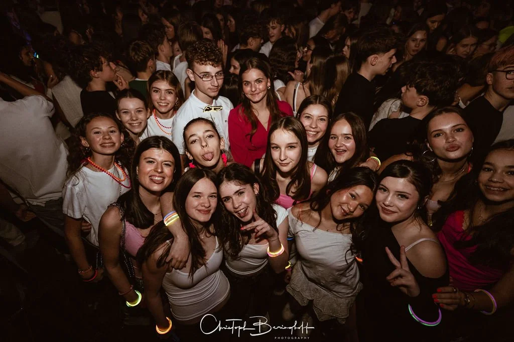 A group of young people at a party; many are smiling, some are wearing neon wristbands, and there is a crowd in the background.