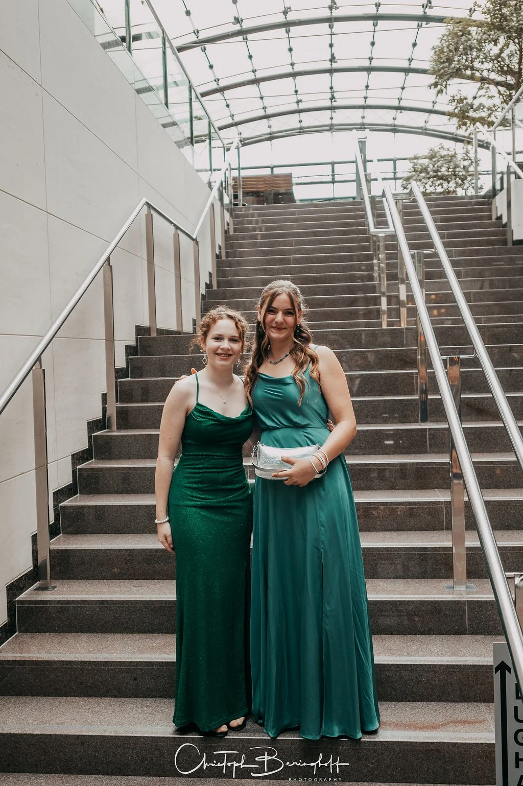 Zwei junge Frauen in eleganten Abendkleidern, die auf einer Treppe stehen, in einem modernen Gebäude mit Glasdach.