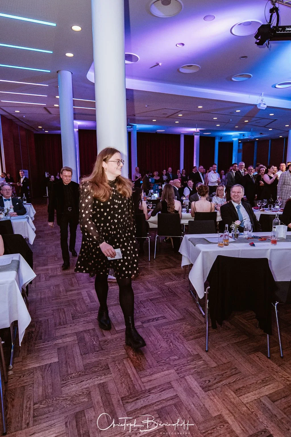 Ein festlich geschmückter Veranstaltungssaal mit vielen eleganten Gästen, die an Tischen sitzen und sich unterhalten, während eine junge Frau in einem schwarzen Kleid mit weißen Punkten und schwarzen Stiefeln in die Kamera lächelt.