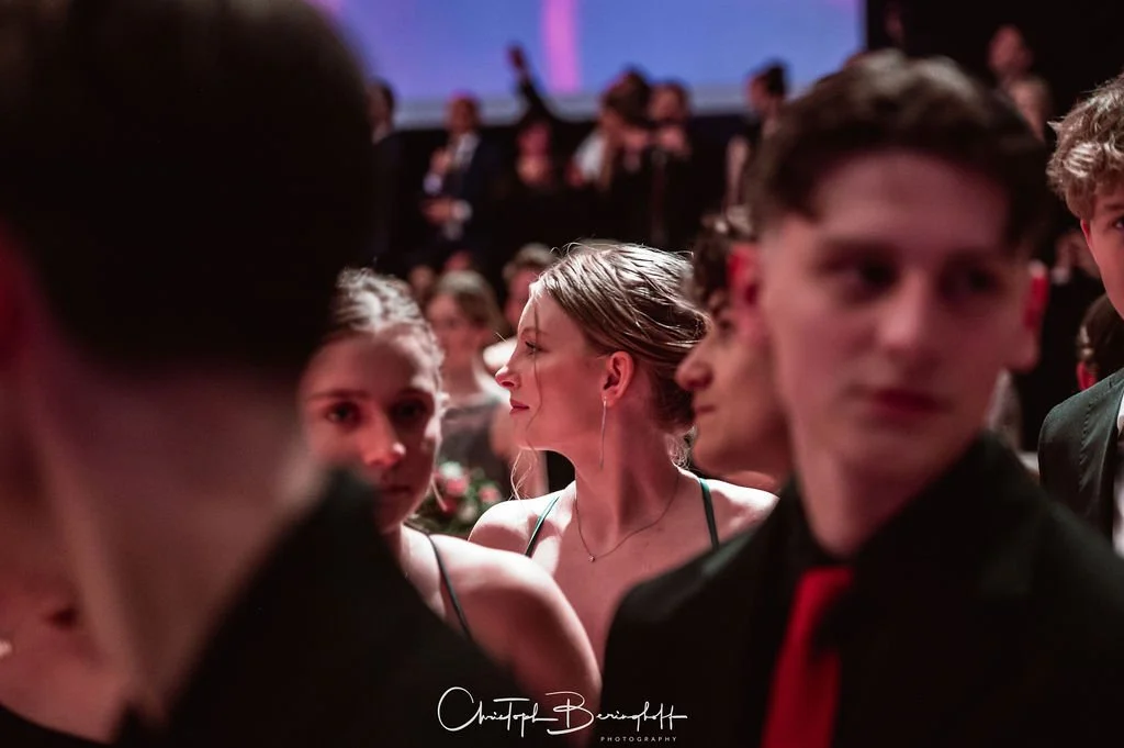 Menschen bei einer formellen Veranstaltung, manche im Anzug oder Abendkleid, im Publikum sitzend, mit einem Fokus auf eine junge Frau mit blonden Haaren in der Mitte.
