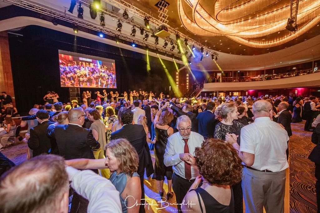 Menschen tanzen und feiern auf einer Ball- oder Festveranstaltung in einem eleganten Saal mit mit Sekt und Musik, große Bühne mit Videoanzeige und Lichtshow, viele Gäste in formeller Kleidung.