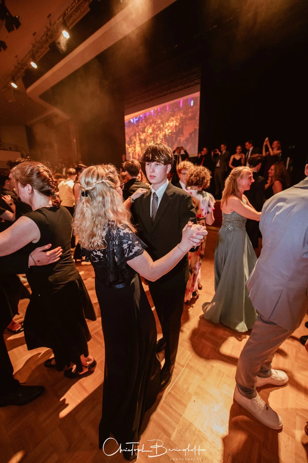 Menschen tanzen auf einer festlichen Veranstaltung im Ballroom, mit einem jungen Mann in dunklem Anzug und einer Frau in schwarzem Kleid im Mittelpunkt, im Hintergrund eine große Leinwand mit hellem Licht.