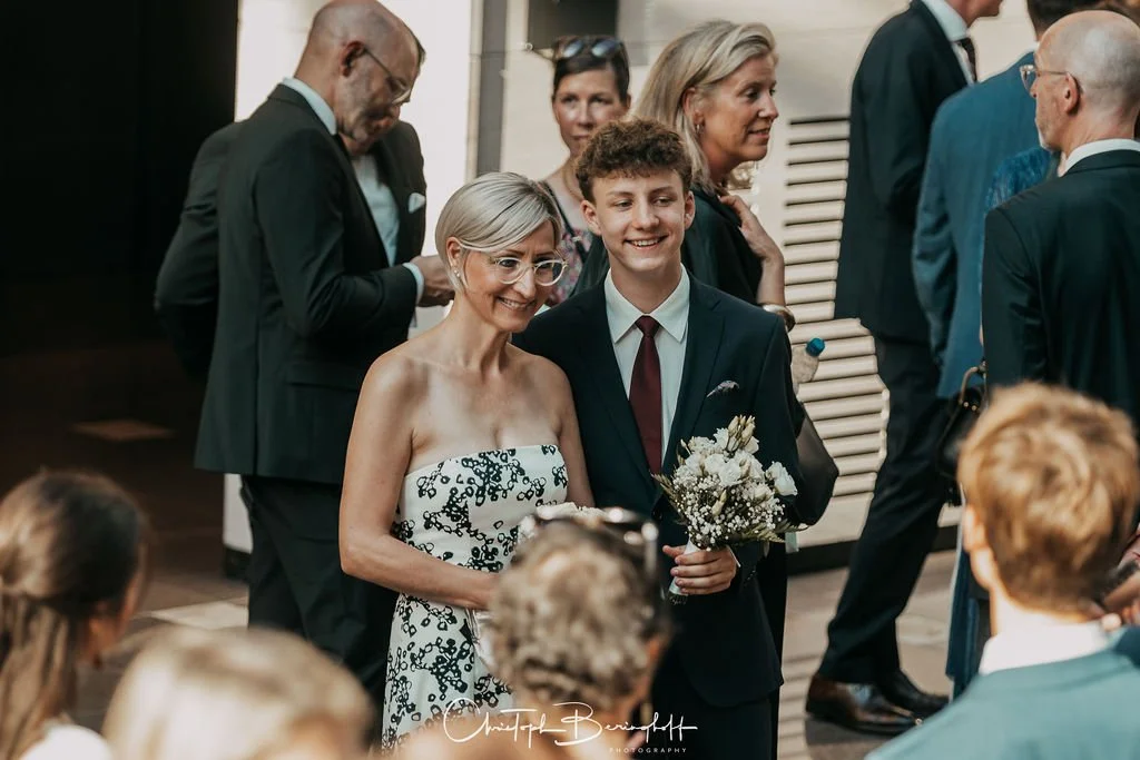 Eine Gruppe von Menschen bei einer formellen Veranstaltung, mit einem lachenden jungen Mann in Anzug, der eine Frau im schwarzen und weißen Kleid hält.