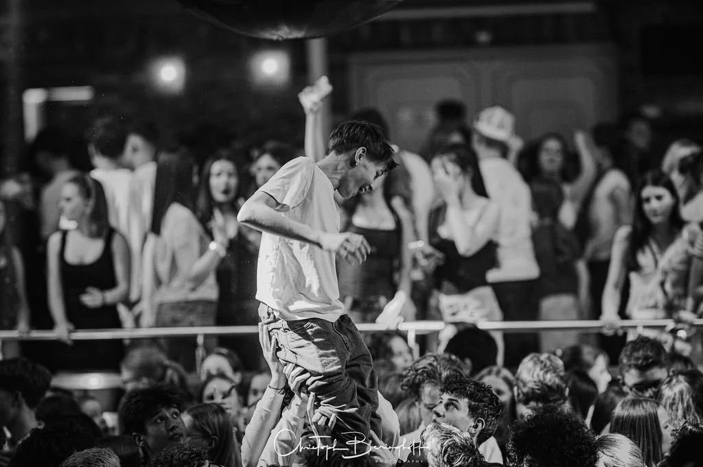 A boy in a school uniform is dancing at a concert with a large crowd in the background.