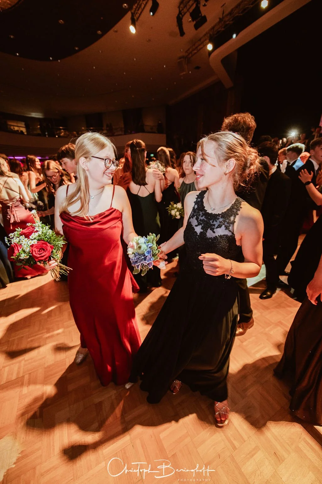Zwei Frauen tanzen auf einer Feier, beide halten Blumensträuße, im Hintergrund viele weitere Menschen auf einem festlichen Tanzboden.