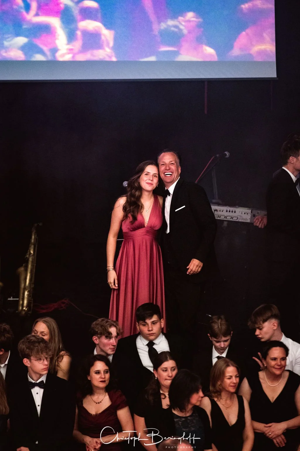 Zwei Menschen in Abendkleidung, ein Mann in Smoking und eine Frau in rotem Abendkleid, posieren an einem Fest oder Event, umgeben von anderen Gästen.