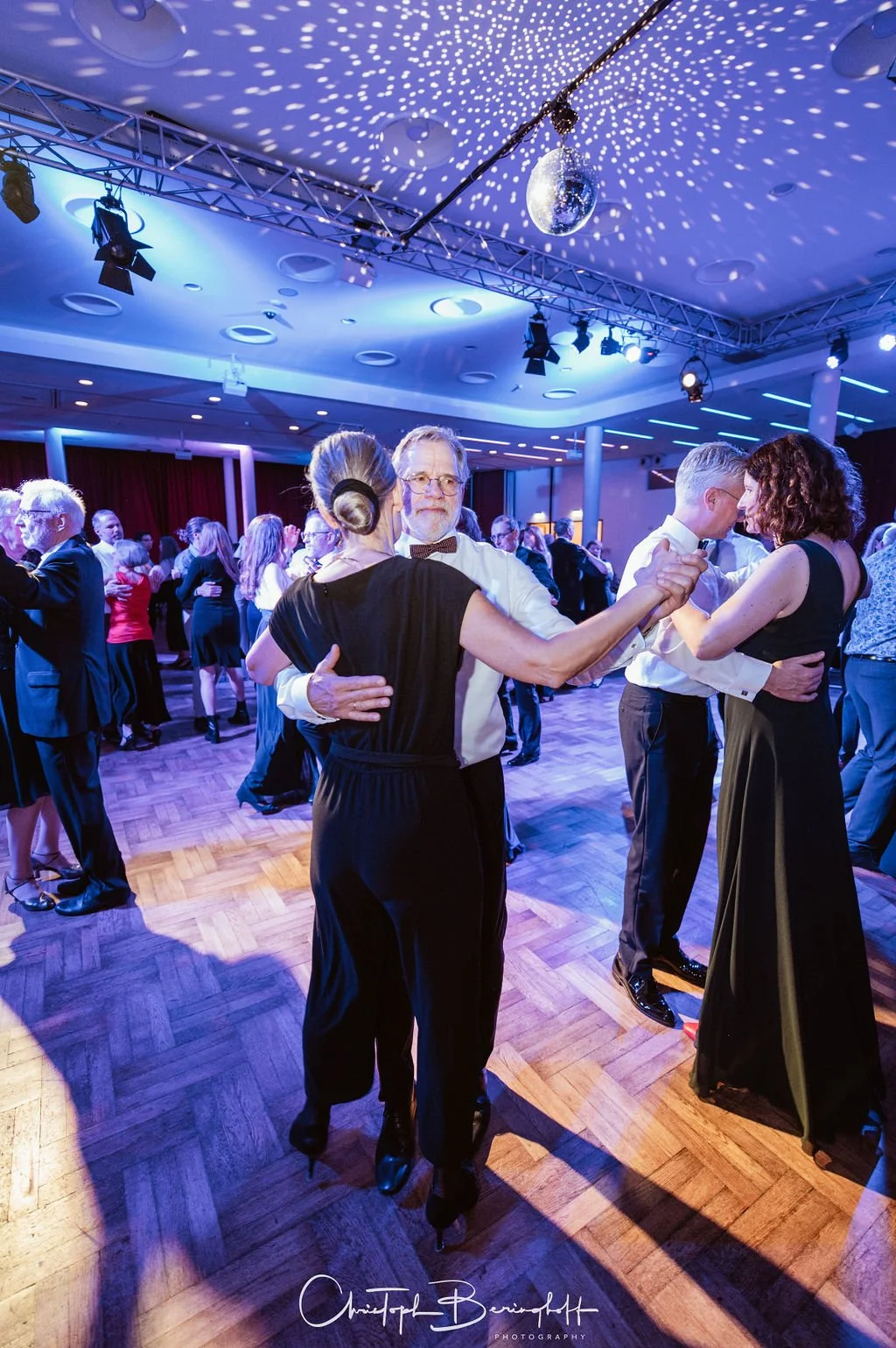 Paare tanzen in einem festlich beleuchteten Saal mit Disco-Kugel und Tanzfläche.