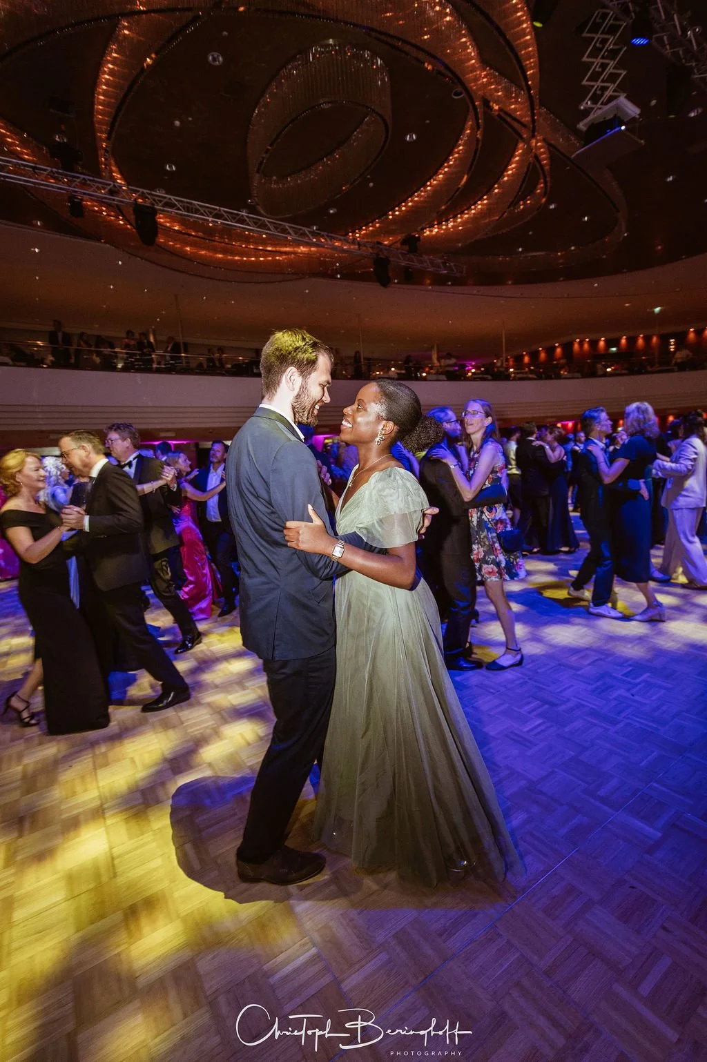 Ein Mann und eine Frau tanzen auf einem festlichen Ball, umgeben von anderen Paaren, in einem großen, eleganten Veranstaltungsraum mit schimmernder Decke und buntem Licht.