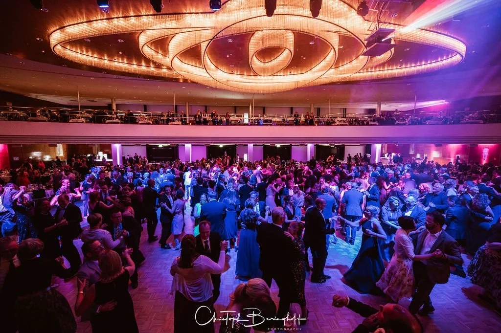 Menschen tanzen auf einer großen Ballsaal-Tanzfläche bei einer Veranstaltung mit buntem Licht. Es sind viele Gäste in eleganter Kleidung. Oben befindet sich eine Galerie mit weiteren Gästen und Tischen, ein bezaubernder, moderner Kronleuchter leuchte