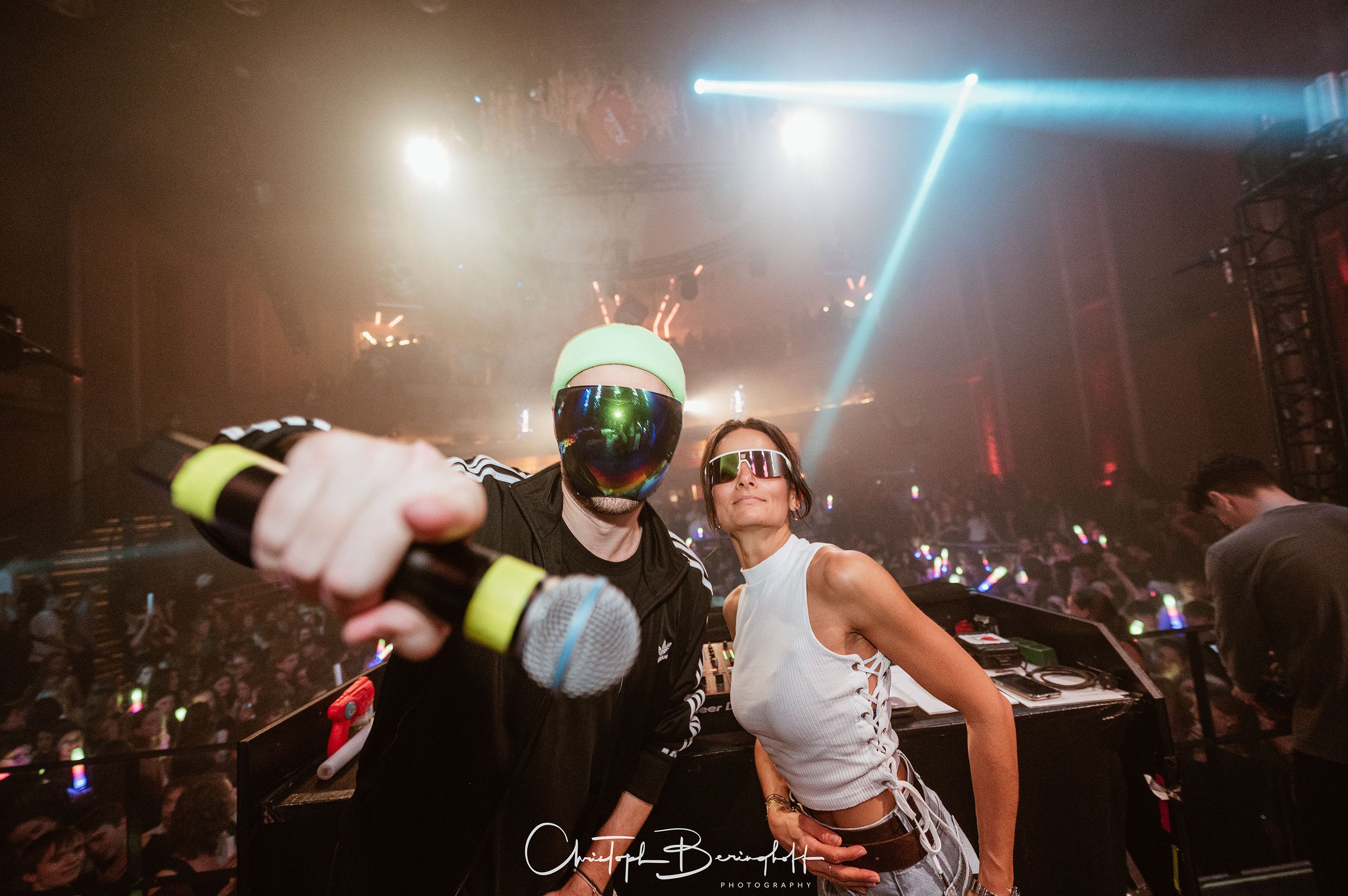 Zwei DJs mit futuristischen Sonnenbrillen und einem Mikrofon in einer dunklen, lichtdurchfluteten Club-Umgebung, umgeben von tanzenden Menschen mit leuchtenden Accessoires.