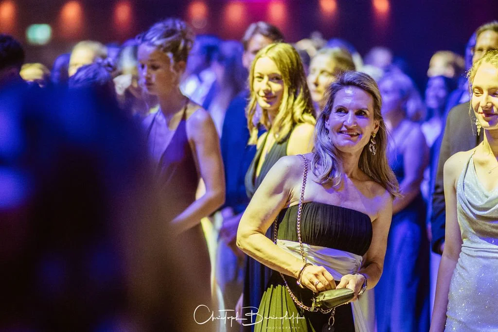 Frauen in eleganten Abendkleidern bei einer Veranstaltung, im Hintergrund bunte Beleuchtung.