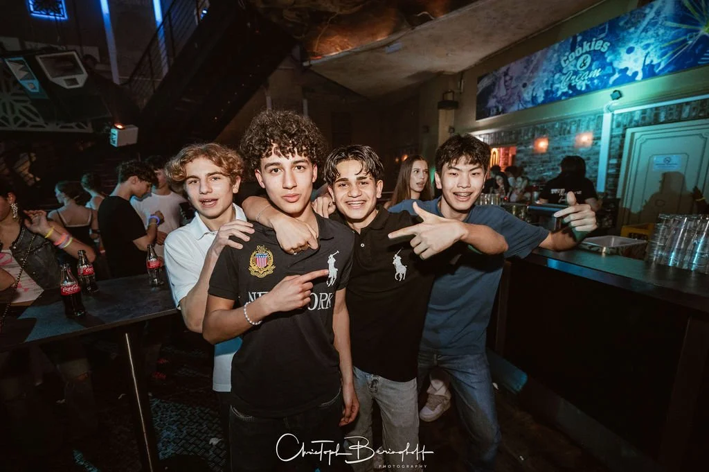 Four teenagers are partying in a bar, with a few bottles of Coca-Cola on the table, colorful lights, and a festive atmosphere.