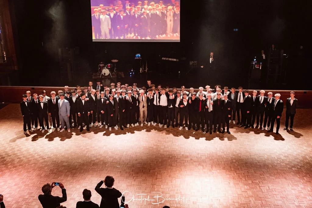 Gruppe junger Männer in formeller Kleidung auf einer Bühne, vermutlich Abschlussfeier oder Konzert, mit einem großen Bildschirm im Hintergrund, auf dem eine ähnliche Gruppe zu sehen ist.