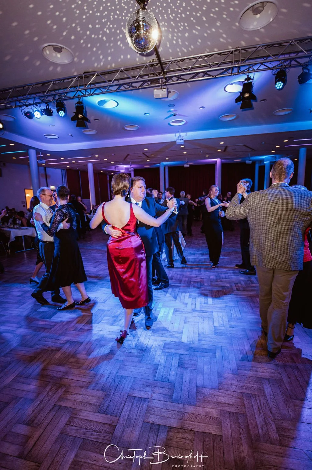Paare tanzen auf einer Tanzfläche in einem festlich beleuchteten Saal, Disco-Kugel an der Decke, elegante Kleidung, Frauen in Kleidern, Männer in Anzügen.