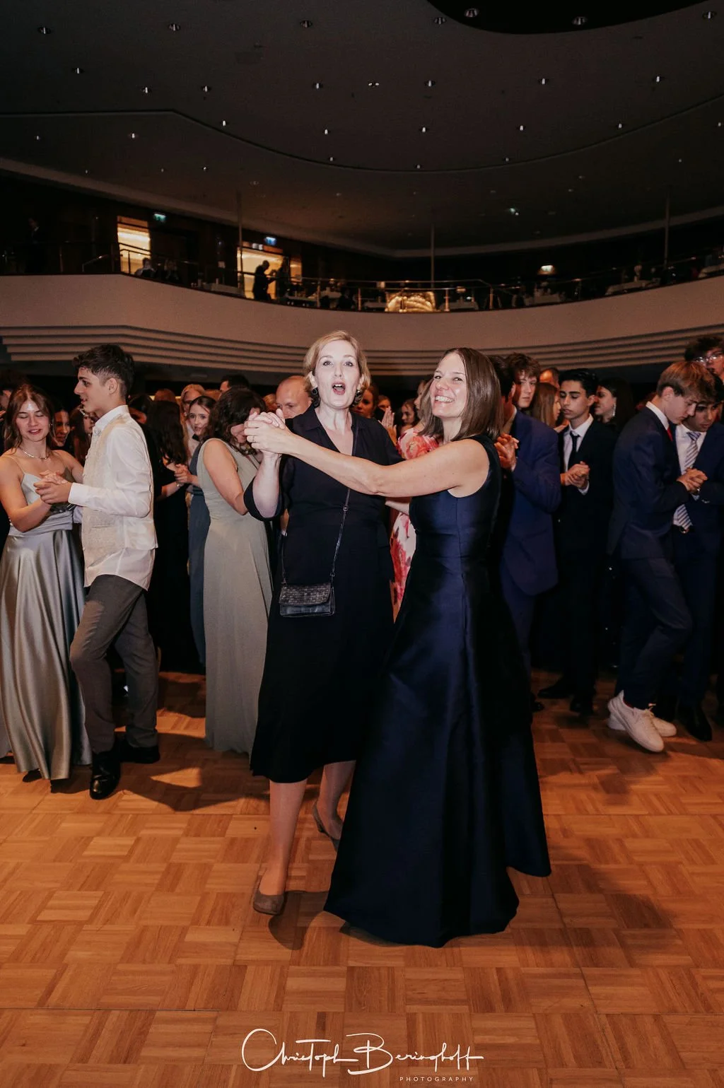 Zwei Frauen tanzen auf einer Feier im dunklen Saal, umgeben von weiteren Gästen in formeller Kleidung.