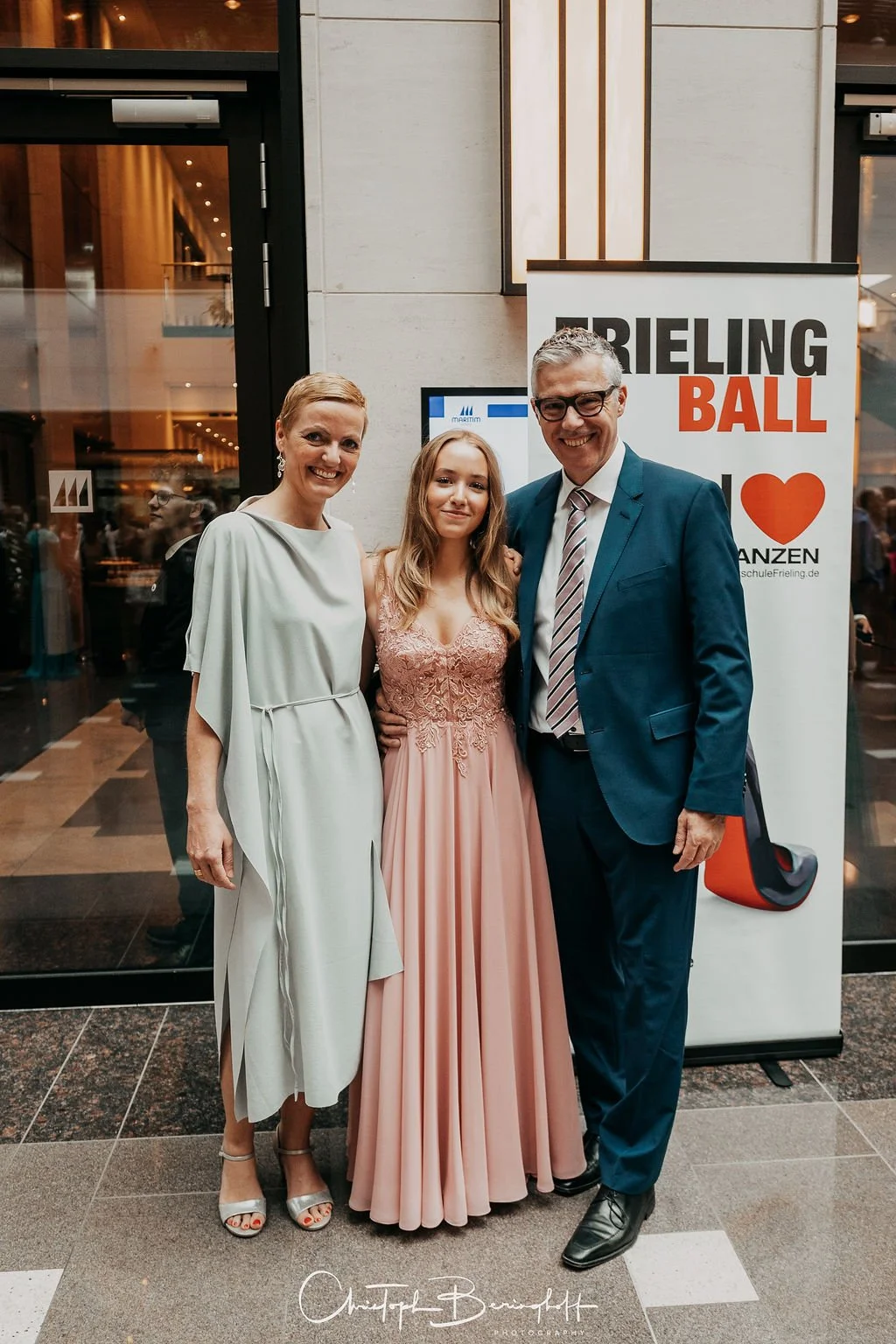 Drei Menschen in formeller Kleidung bei einer Veranstaltung, lachend vor einem Banner mit der Aufschrift 'Brieling Ball' und Herzsymbol.