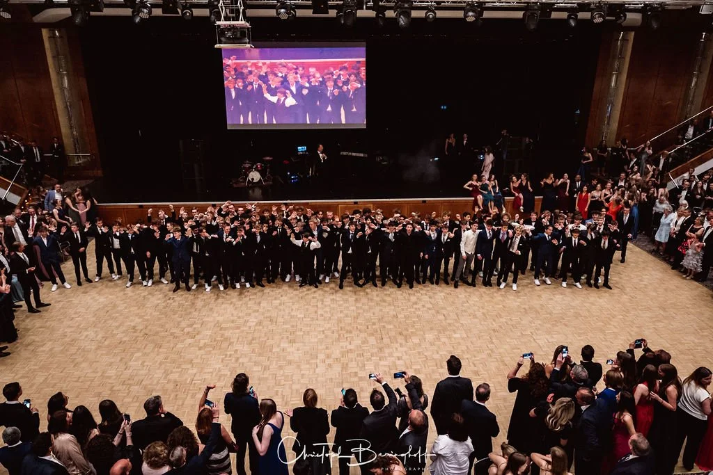 Junge Menschen in formeller Kleidung versammeln sich in einem großen Saal bei einer Feier oder einem Abschluss, während Fotografen und Zuschauer sie aufnehmen.