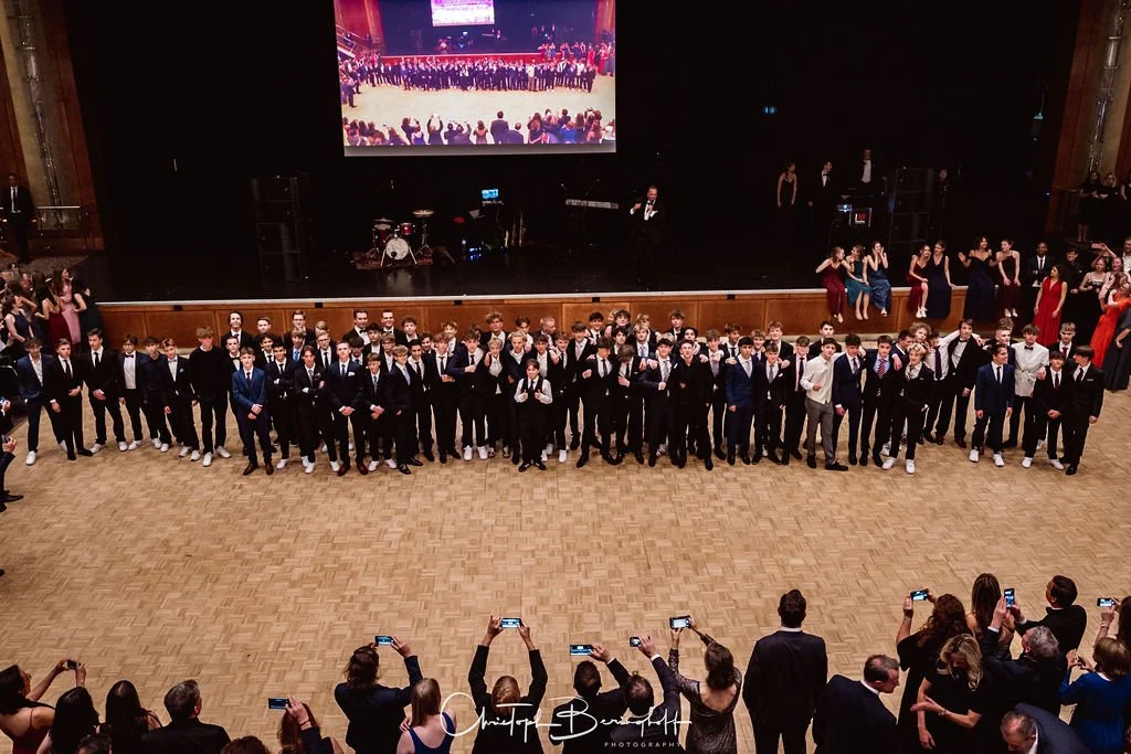 Gruppe junger Männer in Anzügen steht auf der Tanzfläche bei einer formellen Veranstaltung, im Hintergrund eine Bühne mit Musikern, und Personen, die Fotos machen.