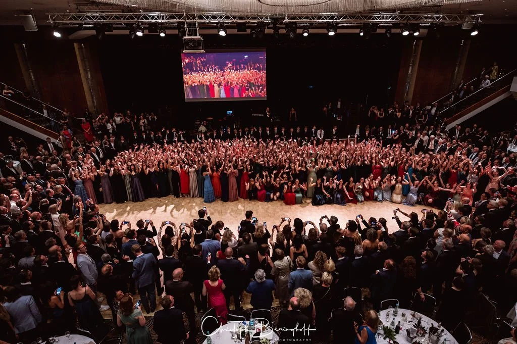 Viele Menschen in formeller Kleidung in einem Festsaal beim Abschlussball, Gruppenfoto auf der Bühne, Zuschauer mit Handys im Publikum