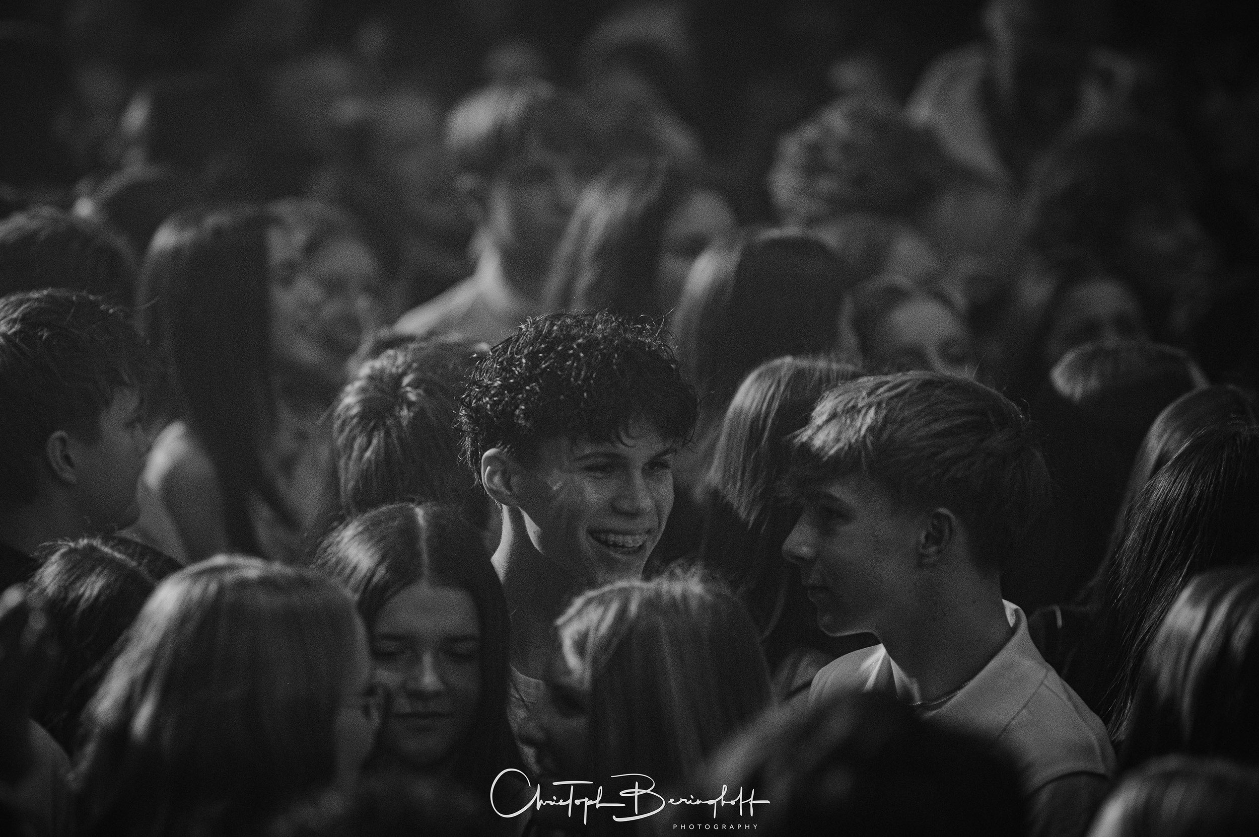 Schwarz-Weiß-Bild von einer Gruppe junger Menschen, die auf einem Konzert oder einer Party tanzen und Spaß haben, im Mittelpunkt ein lachender Junge mit dunklen, lockigen Haaren.