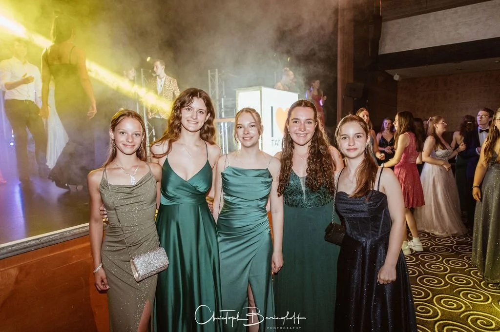 Fünf junge Frauen in eleganten Abendkleidern posieren auf einer Tanzveranstaltung mit Tanzfläche und Musik im Hintergrund.