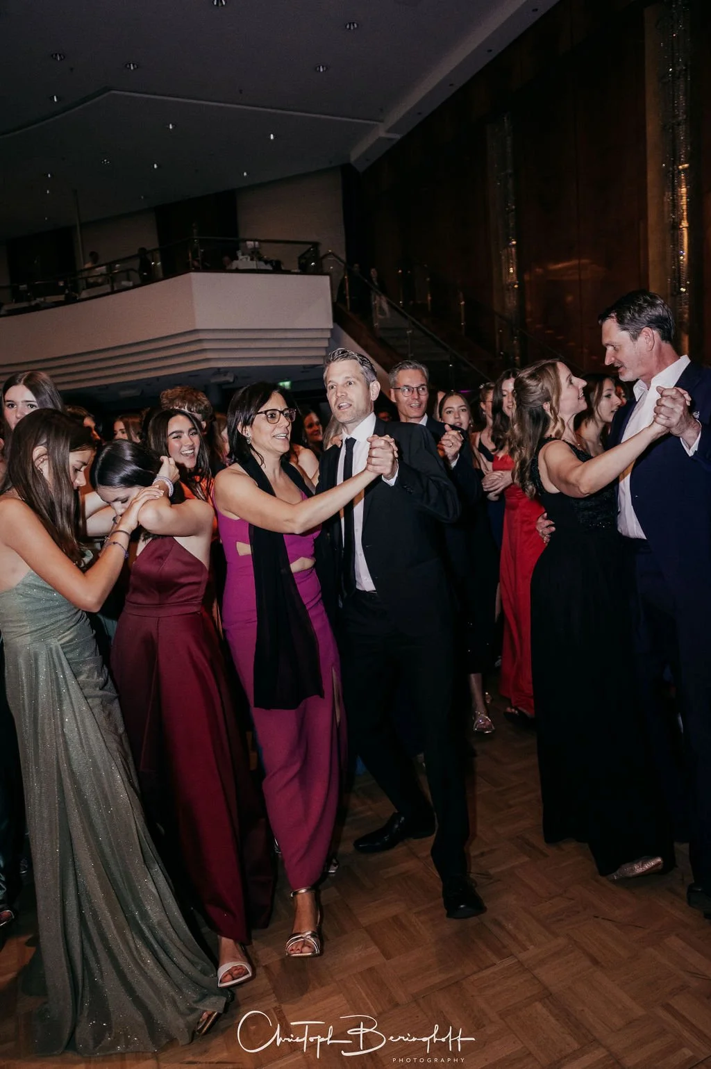 Menschen tanzen bei einer formellen Veranstaltung in einem luxuriösen Saal, viele Frauen in eleganten Abendkleidern und Männer in Anzügen.