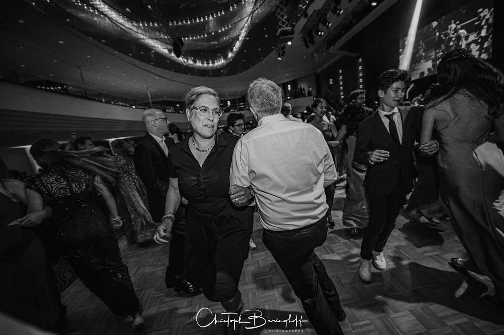 Menschen tanzen auf einer Gala in einem festlich beleuchteten Saal, alle getragen formelle Kleidung, die Stimmung ist lebhaft.