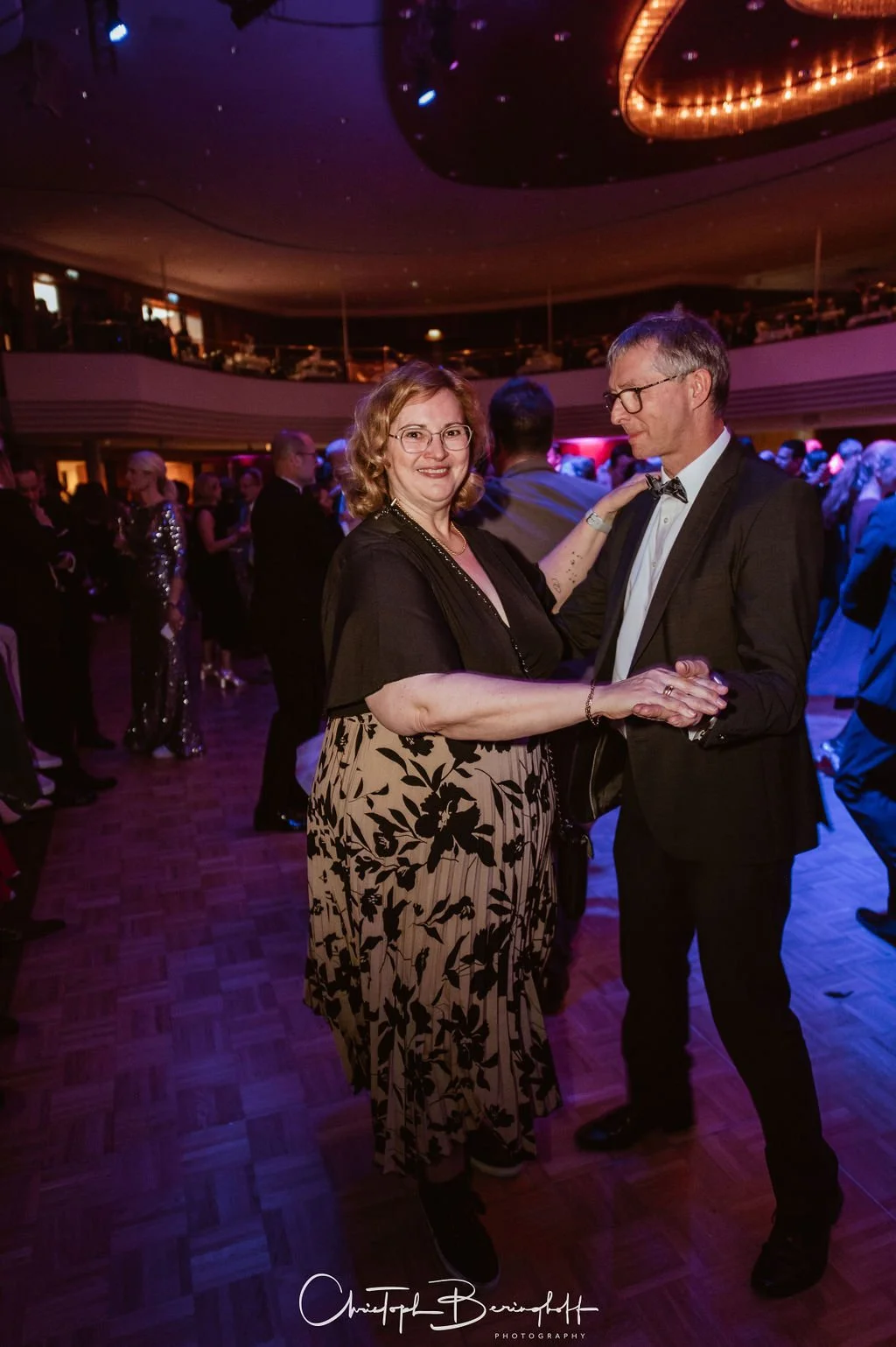 Ein älteres Paar tanzt auf einer festlichen Veranstaltung in einem Ballsaal. Die Frau trägt ein schwarzes Top mit einem floral bedruckten Rock, der Mann einen schwarzen Anzug mit Fliege. Im Hintergrund sind andere Gäste, die auch tanzen, und die Deck