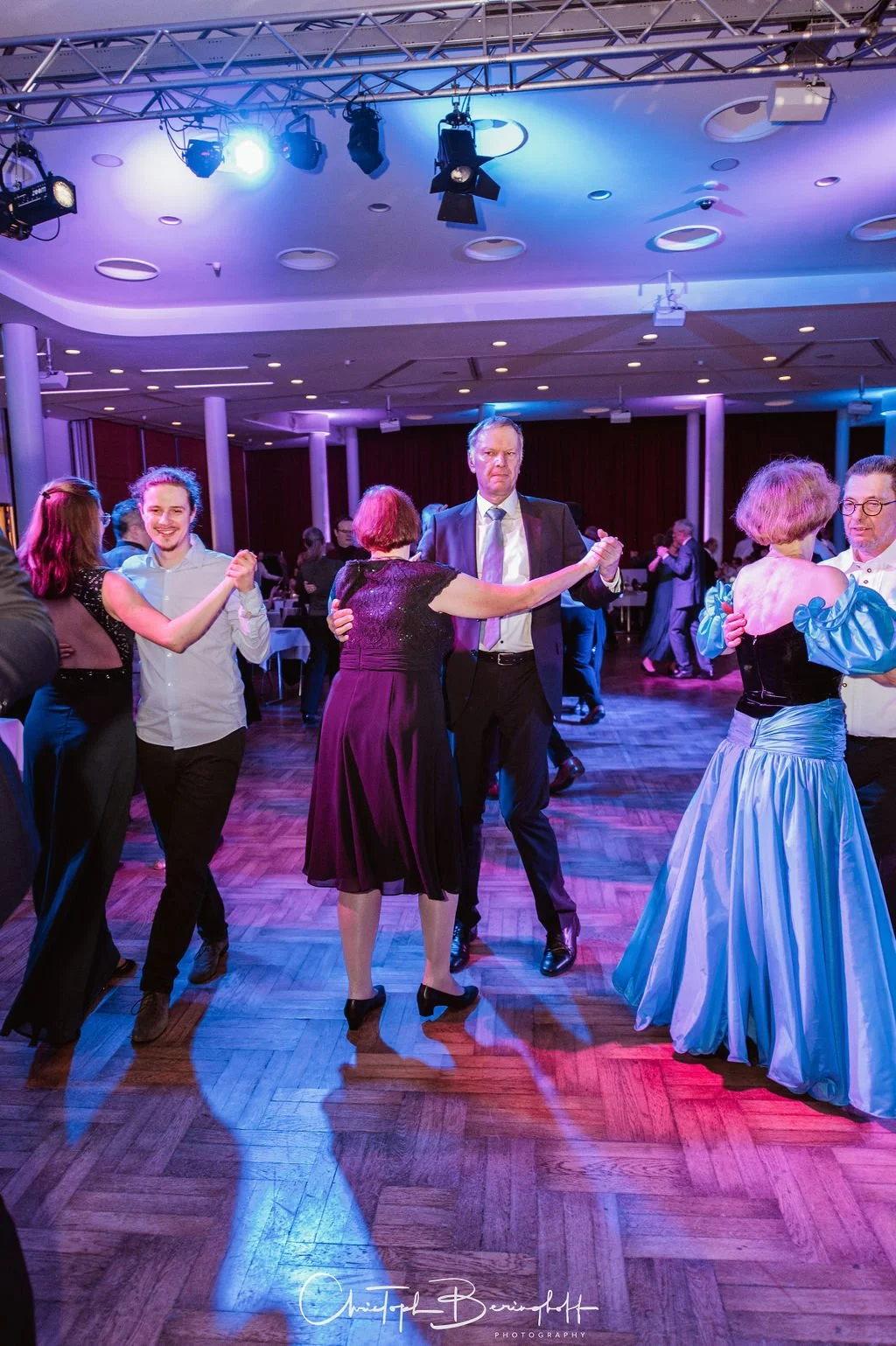 Menschen tanzen auf einer festlichen Tanzfläche bei einer Veranstaltung, einige in elegante Kleidung, mit Lichter und ein Holzparkettboden.
