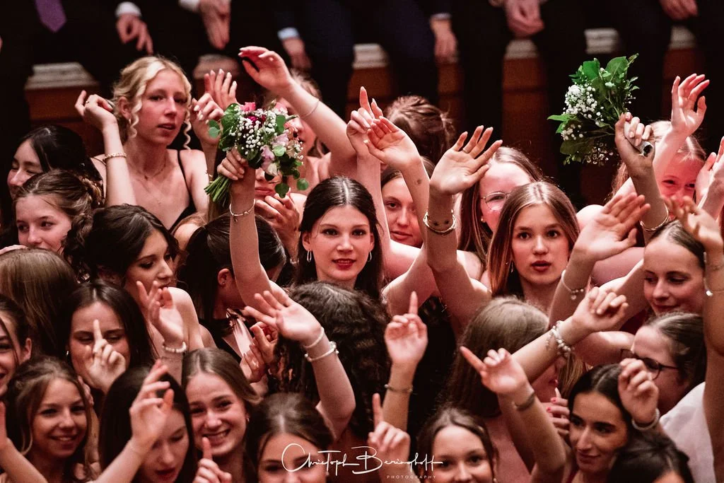 Gruppe junger Frauen bei einer Feier, einige halten Blumen, viele winken und lächeln, im Hintergrund sind weitere Personen in dunkler Kleidung.