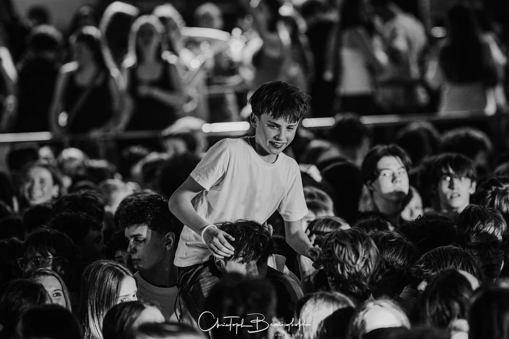 A young boy is sitting on another boy's shoulders in a large crowd, all in a dark, bustling room.