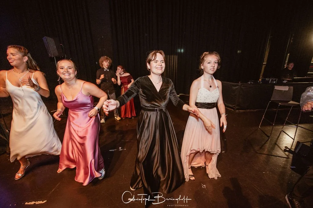 Frauen tanzen auf einer Party oder einem Ball, die meisten tragen elegante Abendkleidung, im Hintergrund schwarze Vorhänge und eine kleine Gruppe weiterer Gäste.