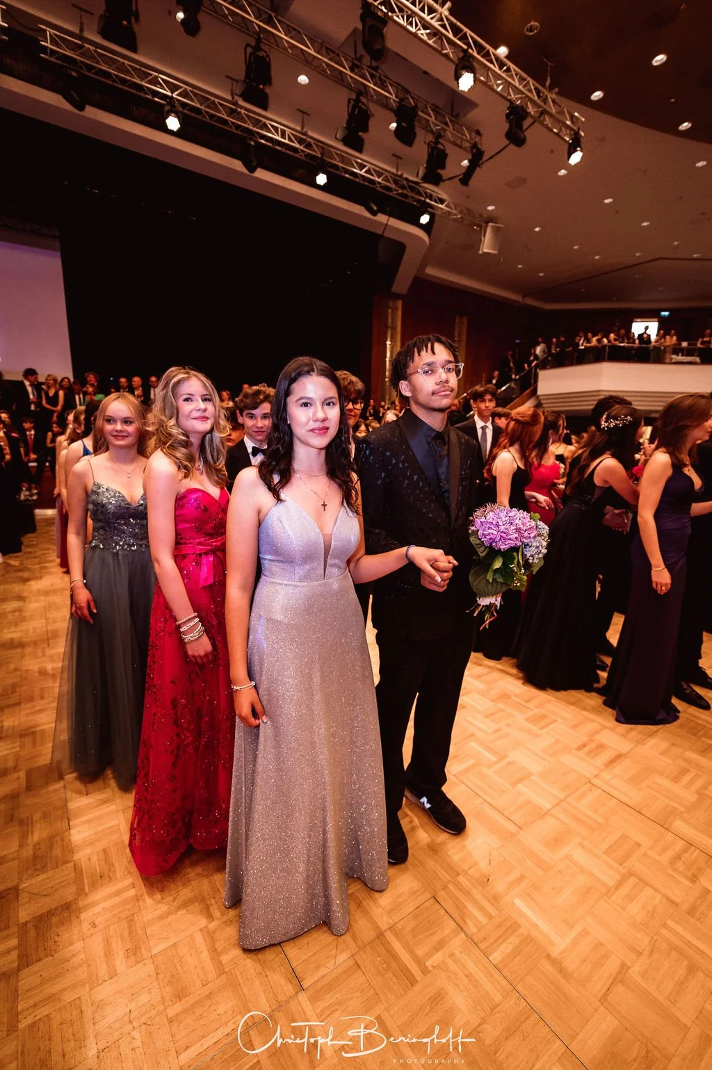 Junge Menschen in festlicher Kleidung bei einer formellen Veranstaltung, einige tragen elegante Abendkleider, während andere Anzüge tragen. Der Fokus liegt auf einer Gruppe, die in einer Reihe auf einem Tanzboden steht, eine Person hält einen Blumens