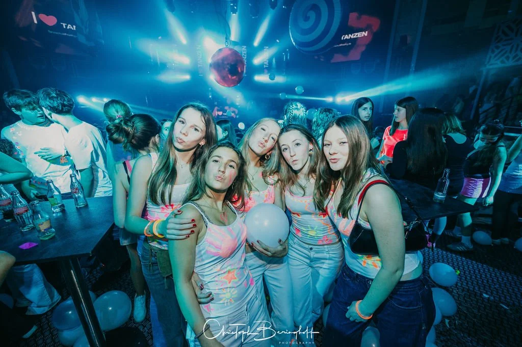 Young women are partying in a nightclub with neon lights and a disco ball; some are striking poses for a group photo, and in the background, more people and drinks are visible.