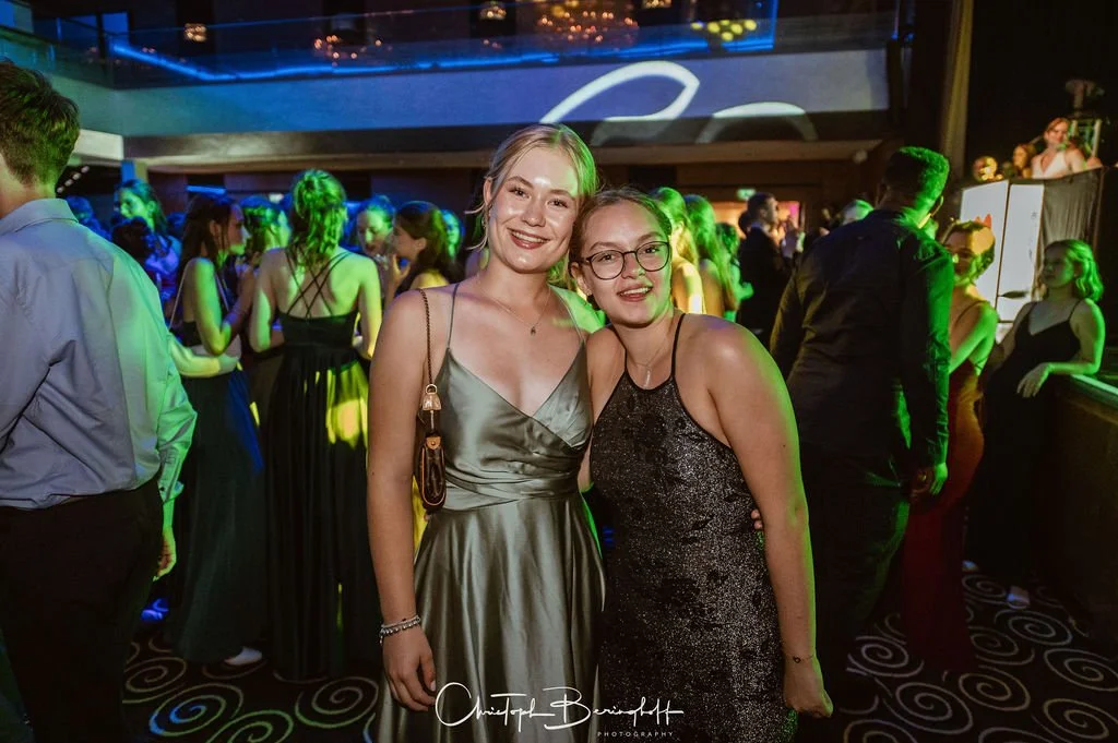 Zwei junge Frauen in eleganten Kleidern auf einer Party, umgeben von anderen Gästen im dunklen Raum mit bunten Lichtern.