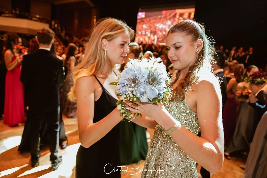 Zwei junge Frauen bei einer Gala oder Ball, eine überreicht der anderen einen Blumenstrauß, im Hintergrund viele weitere Gäste in formeller Bekleidung auf einer festlichen Veranstaltung.